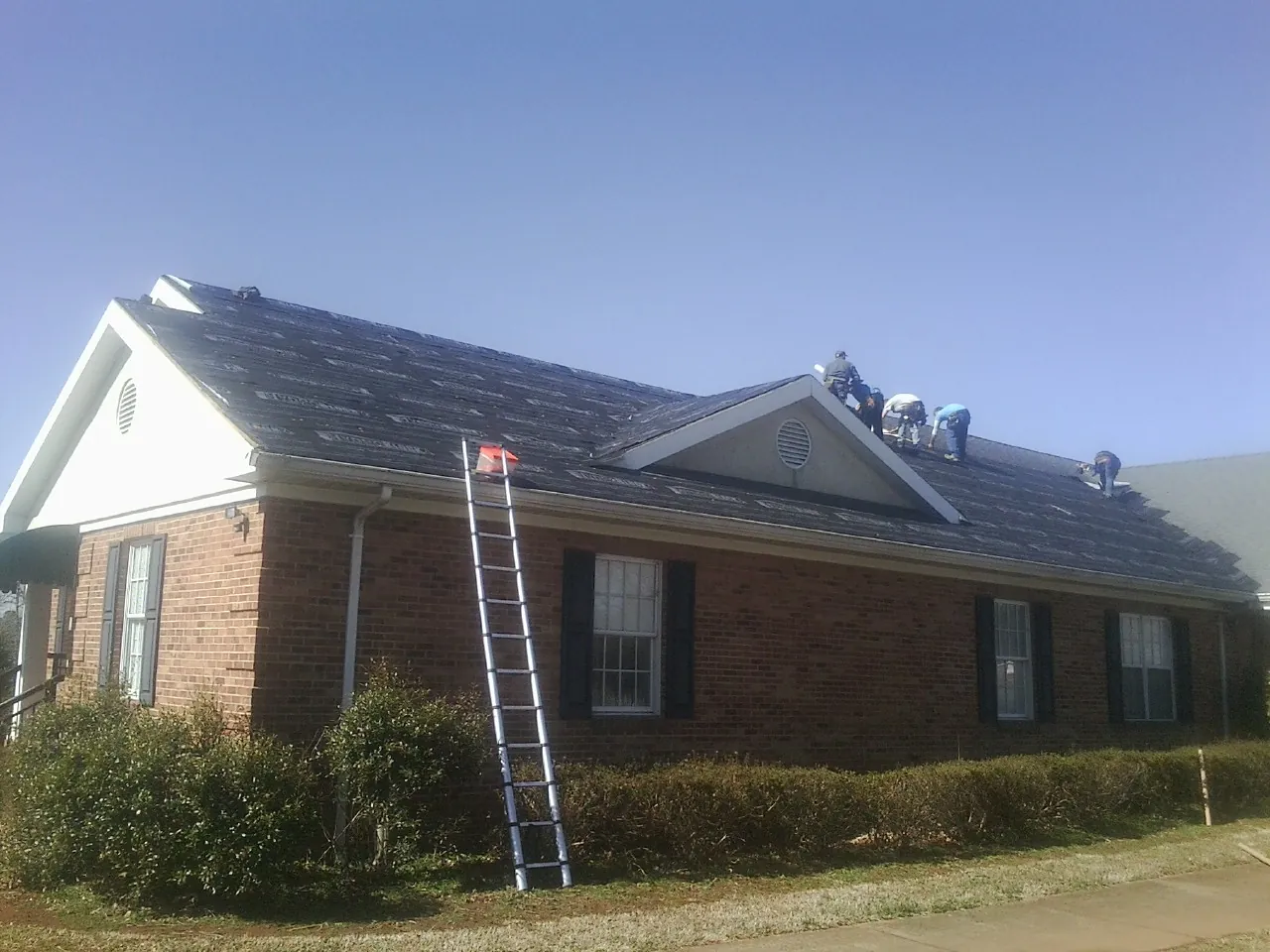 Crew tearing shingles off a brick ranch roof with a ladder set — Metal Roof Repair work by our crew in Pillsbury