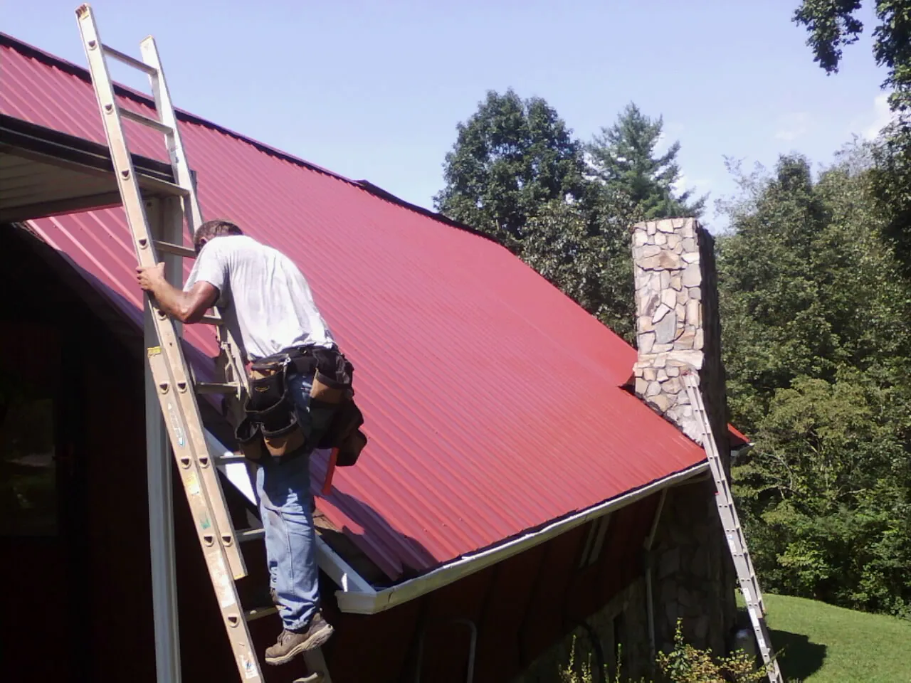 Roofer on a ladder beside a red metal roof and stone chimney — Soffit & Fascia Repair by Roofing Brothers in Pillsbury